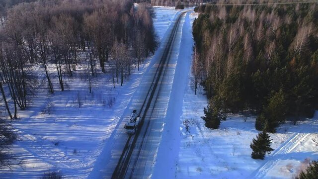 White boom lift truck on empty snowy rural road between forest of coniferous and bare trees at sunset time bird eye view