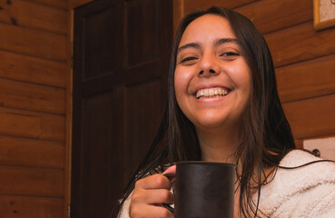 Latina girl relaxing on vacation in a cabin while sipping hot tea from a pewter mug, resting quietly on the bed in her bathrobe.