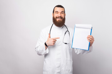 Attractive bearded male doctor in a lab coat displaying a blank clipboard in front of chest and pointing to it.