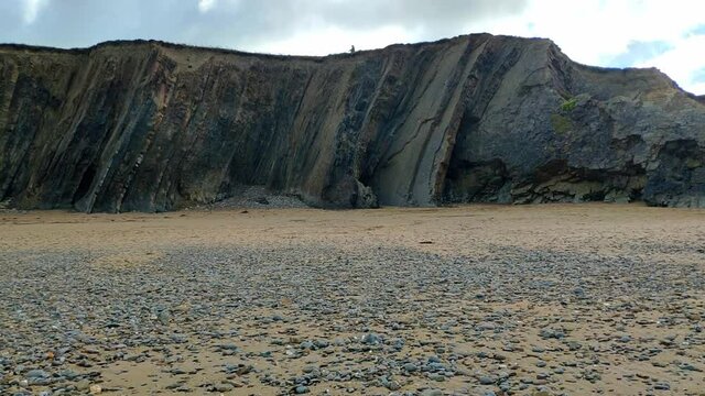 Tall slate rocks aligned vertically forming a cliff a few metres high in Widemouth beach in Conrwall, England. Some people seen walking along the top of it. The beach sand is mostly cover by pebbles.