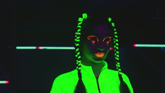 Portrait of female stylish artist inside dark place with neon lights .  Beautiful young girl with fluorescent colorful make-up and cloth in UV light . Portrait of woman under UV black light 