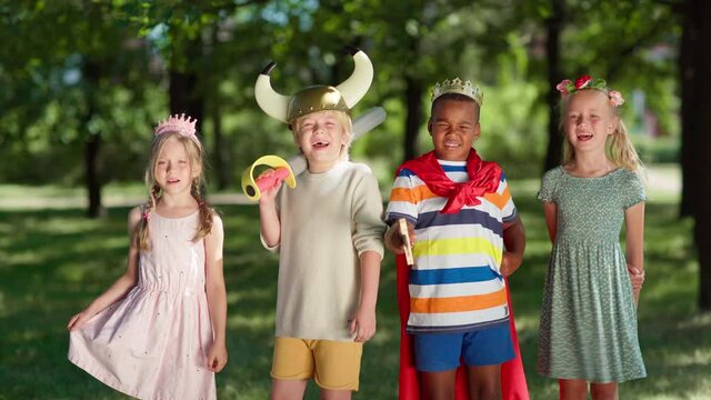 Group of diverse children in costumes looking at camera and screaming joyfully. Boys wearing red cape, king crown and viking helmet, girls in princess diadem and flower headband. Kids role playing