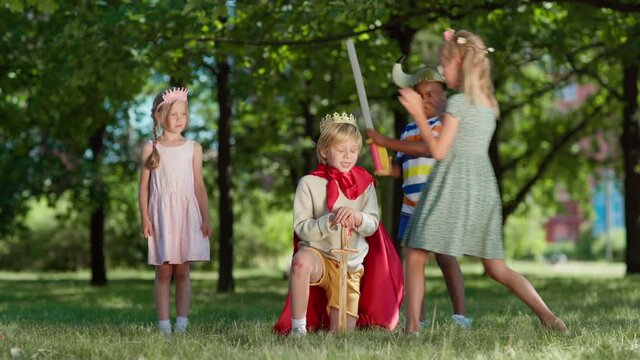 Diverse kids in costumes role playing in park. Little boy in red cape being coronated kneeing on grass with sword in his hand. Ecstatic boy raising arm with sword up. Children applauding to king