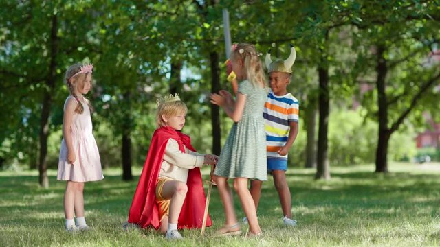 Diverse kids in costumes having fun role playing in park. Little boy in red cape being coronated kneeing on grass with sword in his hand. Boy standing up, crown falling. Children applauding to king