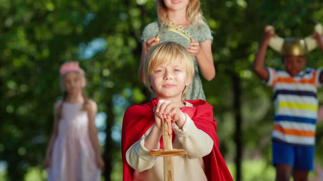 Portrait of kid in red cape and with sword looking at camera role-playing in park. Girl putting crown on boy's head to coronate him. Happy diverse friends clapping hands in background