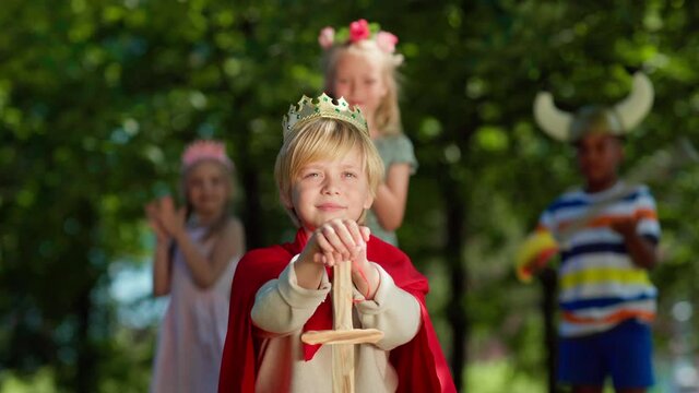 Portrait of confident kid in red cape and with sword looking forwards role-playing in park. Girl putting crown on boy's head to coronate him. Happy diverse friends clapping hands in background
