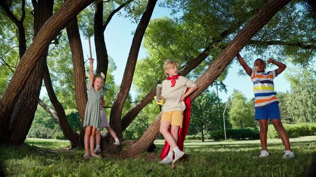 Group of diverse kids in costumes resting by tree while having break in role-playing game in park. Children with toy swords talking and rehearsing further actions in game