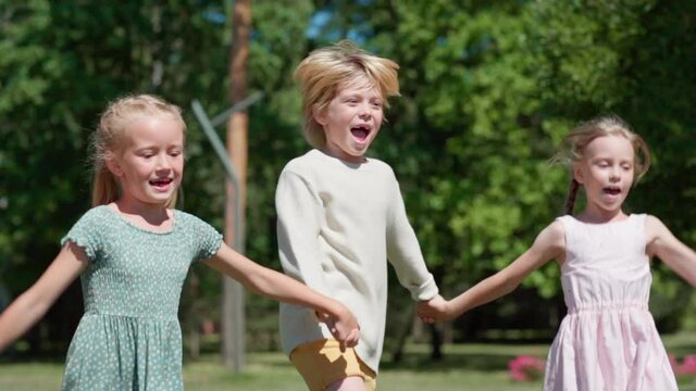 Side view of group of three ecstatic blonde kids, boy and two girls, running holding hands and screaming with excitement. Friendly children hopping in park in slow motion feeling happy and carefree