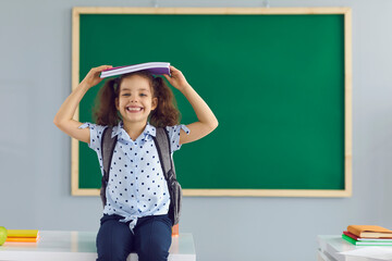 Fototapeta na wymiar Concept back to school. Happy schoolgirl holds a book over her head laughing standing on the background of a green school board in the classroom. Education learning lessons parenting students.