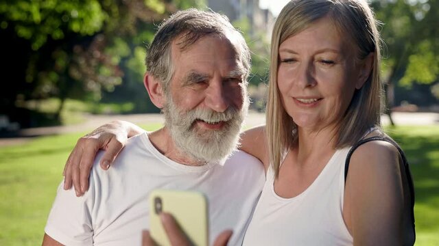 Elderly Man And Woman In The Park After Fitness Talking On Video Call With Someone. Gray Woman Holding Phone In Her Hands.