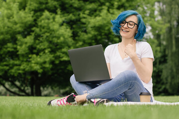 Young women with blue hair sitting on green grass with laptop in the hands.