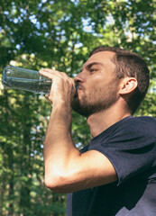 Adult man drinking water from reusable plastic bottle after sport in nature. Recovery, refreshing, healthy lifestyle.