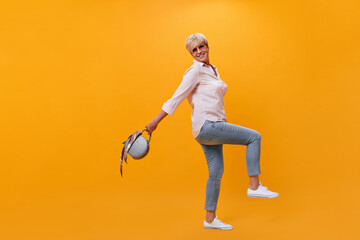 Cheerful lady in street style outfit holds handbag on orange background