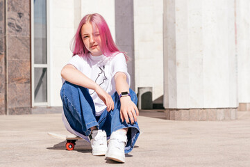 A pink-haired hipster teenage girl in a white T-shirt and jeans is sitting on a skateboard on a...