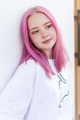 Portrait of a positive pink-haired teenage girl in a white T-shirt leaning against the white wall...