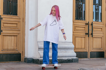 A pink-haired hipster teenage girl in a white T-shirt,jeans rides a skateboard on a city street on a summer day.Generation Z style,active lifestyle.