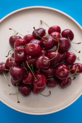 Overhead and closeup view of a ceramic dish full of bright fresh appetizing cherries on blue background.