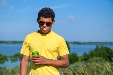 Smiling African American man in sunglasses enjoying the taste of coffee outdoors in summer by the river, looking and standing in front of the camera. Copy space