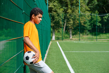 Side view portrait of young African-American man holding soccer ball while standing with back against fence in sports court outdoors, copy space
