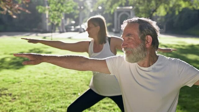 Elderly Man And Woman With Gray Hair Doing Fitness In Park. They Keep Their Arms Outstretched.