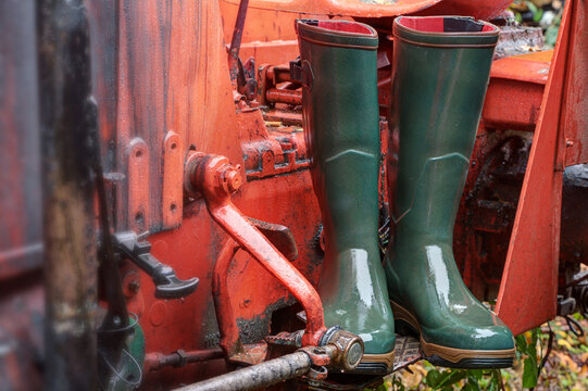 Green rubber boots stand on the step of a bright red tractor in the rain. Wellington boots offer optimal protection on rainy days and are one hundred percent waterproof.