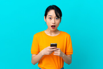 Young Vietnamese woman isolated on blue background looking at the camera while using the mobile with surprised expression