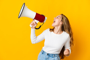 Young blonde woman isolated on yellow background shouting through a megaphone to announce something...