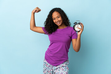 Teenager cuban girl isolated on blue background in pajamas and holding clock while doing strong gesture