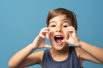 Front view of small caucasian boy four years old standing in front of blue background studio shot standing and holding hands in front of his opened mouth like he is shouting