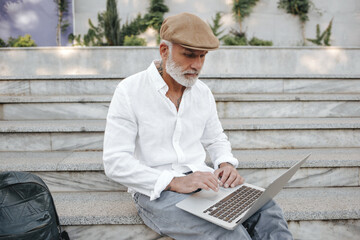 Man in velvet cap works in laptop outside. Serious adult guy with white beard in tattoos and business clothes is sitting on bench..