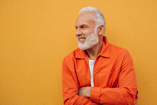 Charming man in orange jacket smiling on isolated background. Gray-haired adult guy with beard in stylish bright clothes with tattoos is smiling..