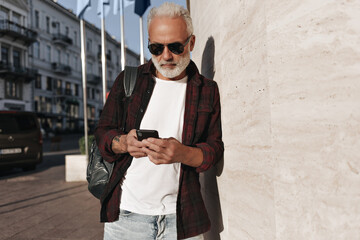 Handsome man leans on wall and chatting in phone. Serious gray-haired guy in burgundy shirt and light T-shirt posing with backpack and smartphone..