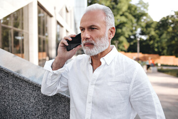 White haired man in light shirt talking on phone. Business attractive adult in stylish clothes posing and holding smartphone..