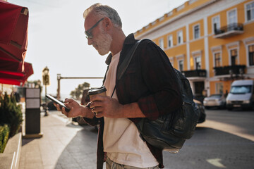 Trendy man in stylish sunglasses, plaid burgundy shirt and white T-shirt chatting on smartphone and holding coffee cup in city..