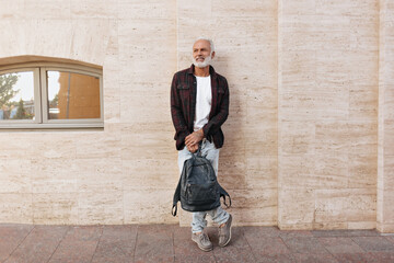 Fototapeta na wymiar Man in jeans and shirt leans on wall and holds backpack. Gray-haired adult fashionable guy in modern clothes smiling on street..