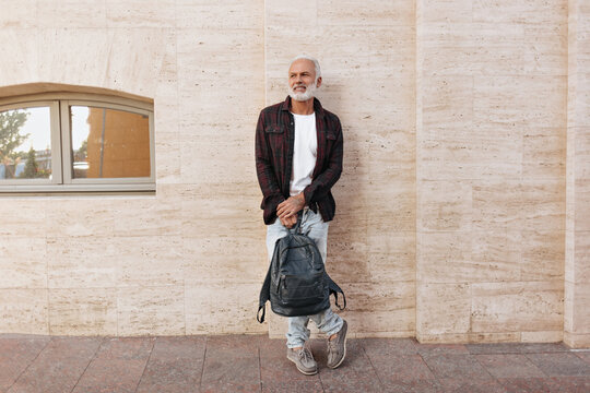 Man in jeans and shirt leans on wall and holds backpack. Gray-haired adult fashionable guy in modern clothes smiling on street..