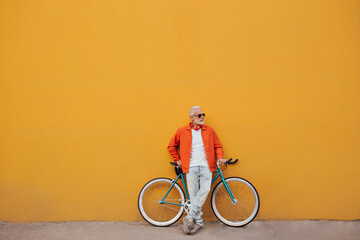 Stylish man in sunglasses leans on bicycle on orange wall background. Gray-haired fashionable guy...