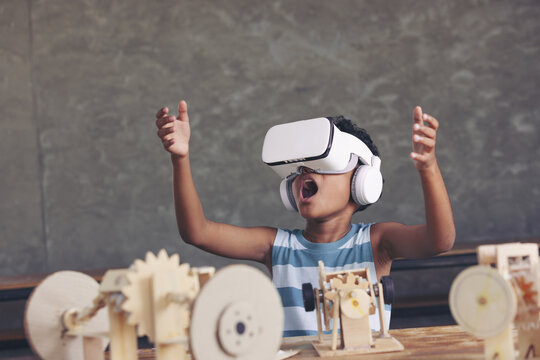 African black boy wearing 3D gadget and looking out above and having the simulation mechanism robot model wooden on table in classroom.