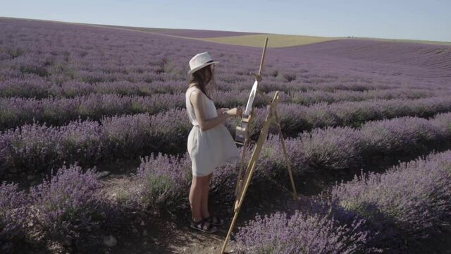 Plein air in nature in a lavender field. Hobbies, art therapy and entertainment. A beautiful young artist girl in a dress and hat draws a picture with paints on a round canvas