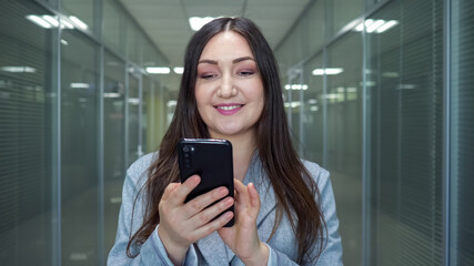 Obraz premium Successful lady company worker looks into smartphone display typing and smiling in office building hallway under electric light in evening closeup