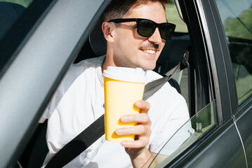 Portrait of a joyful male driver in glasses with a disposable cup of coffee while driving in a car. Happy smiling man driving a car and looking out the open window on a sunny day