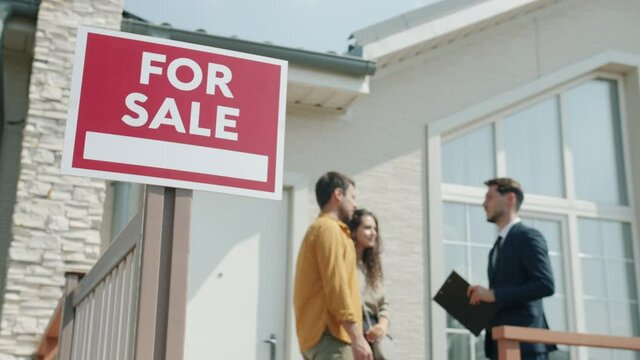 Family man and woman are talking to realtor standing outdoors near house with for sale sign discussing deal and looking at building and lawn. Business and housing concept.