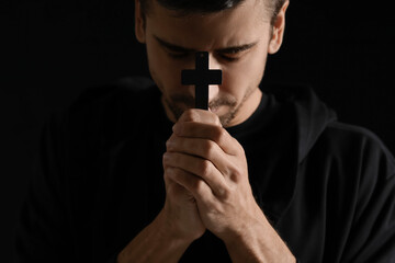 Religious young man with cross praying on dark background, closeup