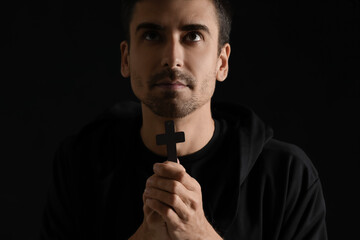 Religious young man with cross praying on dark background