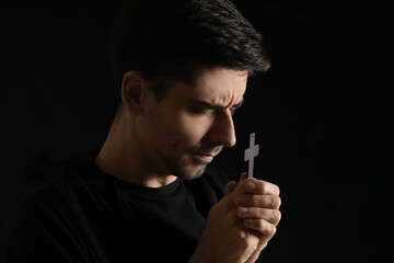 Religious young man with cross praying on dark background