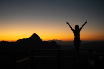 Silhouette of young Asian woman standing on a bench with raising two fingers on the sky while admires the fantastic landscape of mountain peaks with a soft golden horizon line on sunset or sunrise