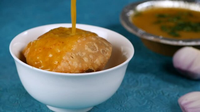Mouthwatering potato gravy falling on Khasta Moong Dal Ki Kachori in a bowl - Morning breakfast from North India. Closeup shot of a bowl of Aloo Ki Sabzi  onion slices - Indian cuisine