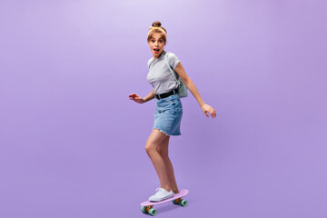 Pretty girl in denim skirt rides longboard. Wonderful cool young woman in grey trendy shirt and white sneakers posing on isolated background..
