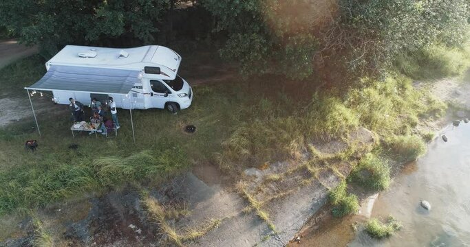 Summertime, group of young people are enjoying a party in nature near the lake, people communicate and relaxing in nature near the camping car, sunset, view from height.