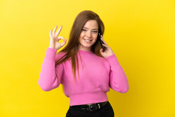 Teenager girl using mobile phone over isolated yellow background showing ok sign with fingers
